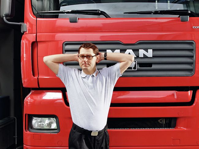 Un homme se trouve devant un camion. Ses deux bras sont levés au niveau des oreilles, ses doigts sont croisés derrière la nuque. L’homme tourne son buste vers la droite.
