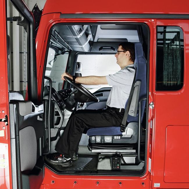 Un uomo siede al volante di un camion. Siede diritto sul sedile e preme la nuca contro il poggiatesta. Le braccia si riposano rilassate sul volante, le spalle sono abbassate.