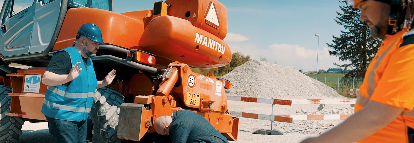 Un homme en tenue de ville se penche sous un gros engin de chantier. Il a enlevé son casque moto. Le temporaire ne semble pas avoir été instruit correctement.