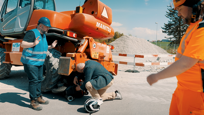Un homme en tenue de ville se penche sous un gros engin de chantier. Il a enlevé son casque moto. Le temporaire ne semble pas avoir été instruit correctement.