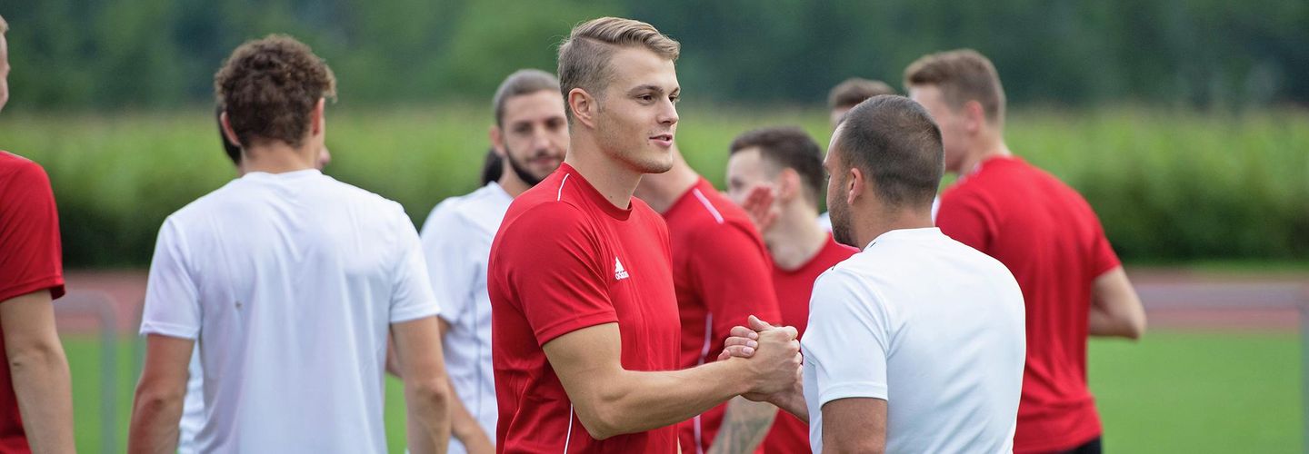 Sportler in weissen und roten Trikots stehen auf einer Rasenfläche. Im Vordergrund reichen sich zwei Männer aus der jeweils gegnerischen Mannschaft die Hand.