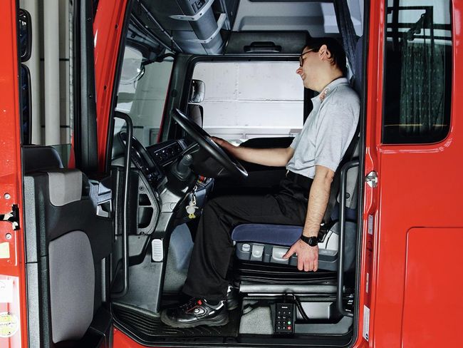 Un uomo siede al volante di un camion. Con la mano destra si tiene rilassato al volante, con la mano sinistra al sedile. Guarda in avanti e piega la testa verso destra.