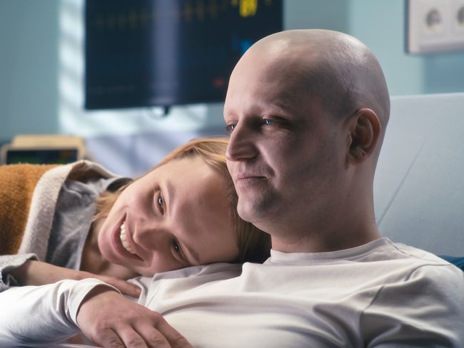 Krebskranker Mann im Bett, der gemeinsam mit seiner Frau etwas auf dem Laptop schaut.