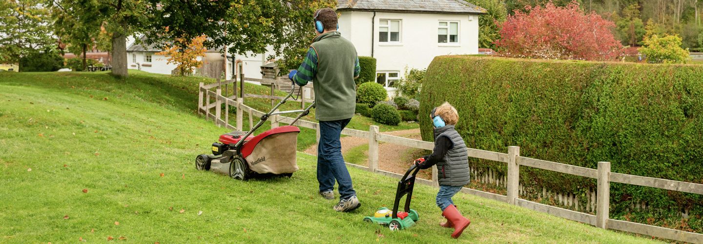 Zuhause, Garten, Rasenmähen, Vater und Sohn, Gehörschutz, Familienzeit, Gartenarbeit, Sicherheit, Outdoor, Wochenendaktivität, Rasenpflege, Natur, Geräuschschutz, Hausarbeit, Eltern-Kind-Moment.