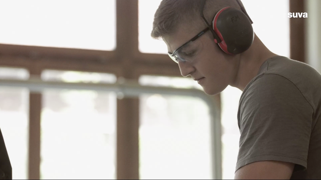 Still aus dem Video. Ein Lernender für den Beruf Schreiner schneidet einen Balken zu. Dabei trägt er eine Schutzbrille und Gehörschutz.
