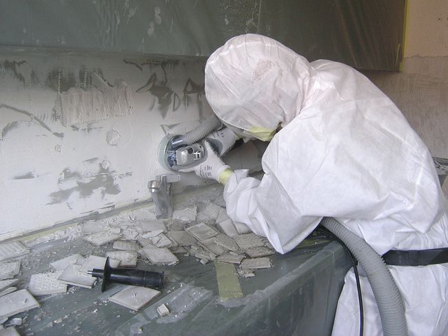 Ein Asbestsanierer entfernt mit einem Schleifgerät den asbesthaltigen Plattenkleber. 