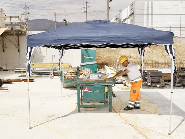 Un operaio in abbigliamento protettivo arancione con elmetto giallo taglia del legno con una macchina da taglio protetta da una tettoia.