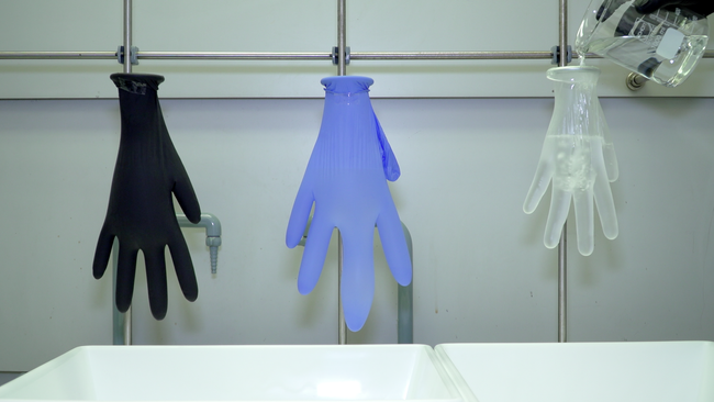 An einer Metallstange hängen ein schwarzer, ein blauer und ein durchsichtiger Handschuh, in den gerade eine Flüssigkeit gegossen wird.