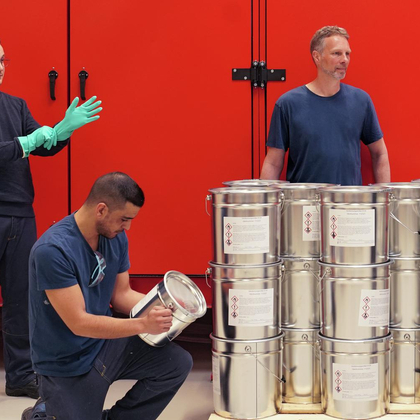 Vier Mitarbeitende stehen um eine Pallete Eimern herum. Auf den Eimern sind verschiedene Warnzeichen zu sehen.