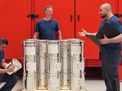 Vier Mitarbeitende stehen um eine Pallete Eimern herum. Auf den Eimern sind verschiedene Warnzeichen zu sehen.