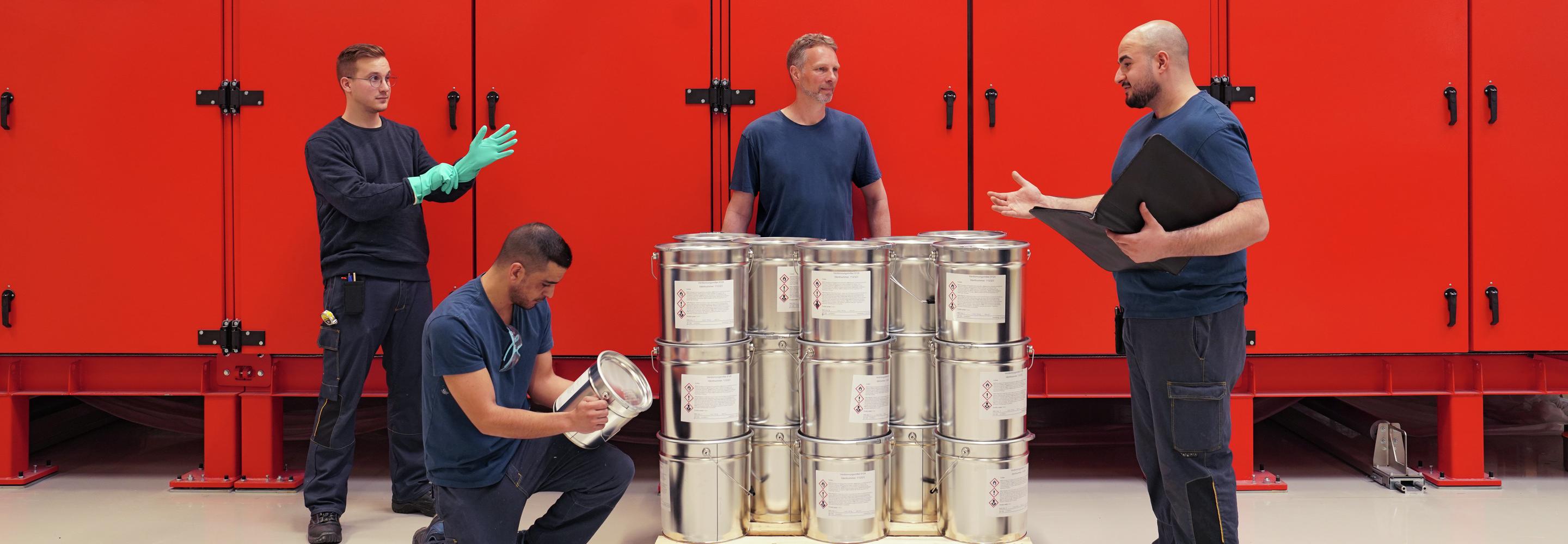 Vier Mitarbeitende stehen um eine Pallete Eimern herum. Auf den Eimern sind verschiedene Warnzeichen zu sehen.