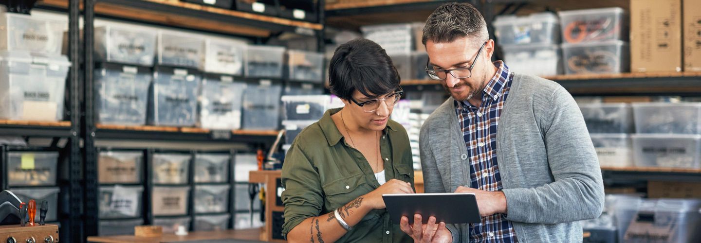 Eine Frau und ein Mann stehen in einer Werkstatt und schauen gemeinsam auf ein digitales Tablet, das der Mann hält. Die Frau zeigt auf das Display, während sie sich über die angezeigten Informationen austauschen.