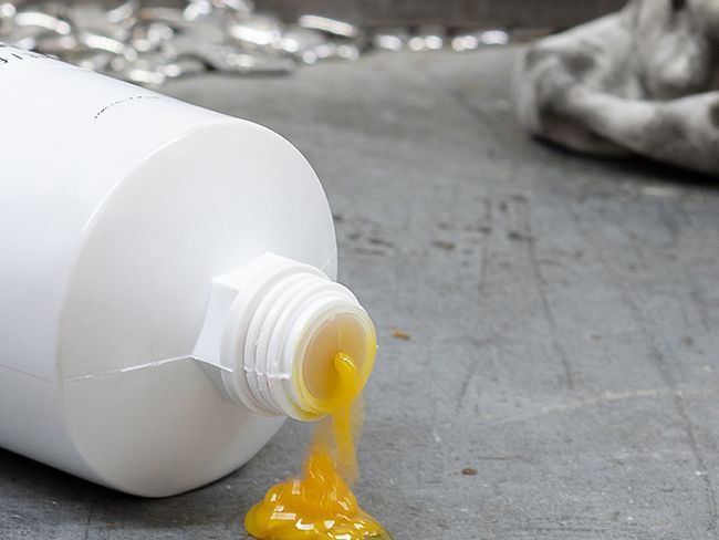 Une substance grasse jaune s’écoule depuis l’ouverture d’une bouteille en plastique blanche sur un sol en béton.