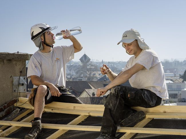Deux couvreurs portant un pantalon long noir, un t-shirt blanc et un couvre-chef blanc doté d’une visière et d’un protège-nuque sont assis sur une charpente de toit. La personne à gauche boit une bouteille d’eau.