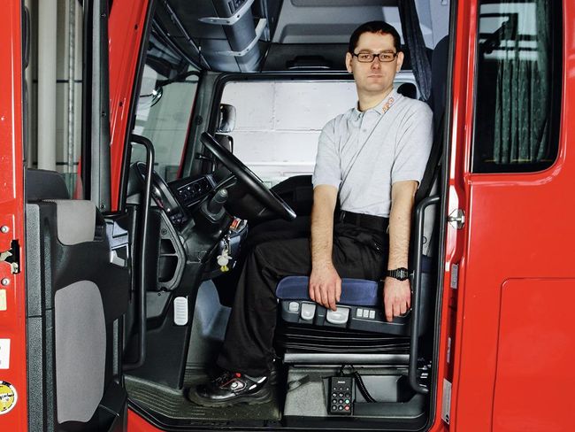 Un homme est assis au volant d’un camion. Les jambes devant lui, il tourne le buste avec les deux bras abaissés sur le côté gauche.