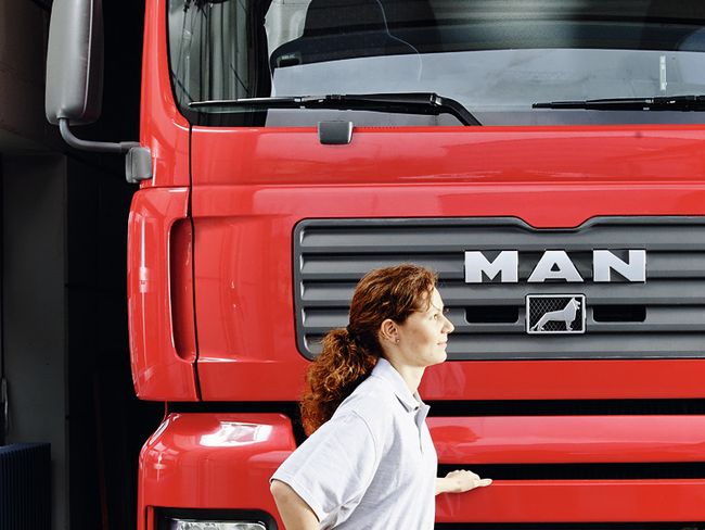 Eine Frau steht seitlich vor einem LKW. Sie stützt sich mit der linken Hand leicht am Fahrzeug ab, hat die rechte Hand in die Taille gestützt und schwingt das rechte Bein nach hinten.