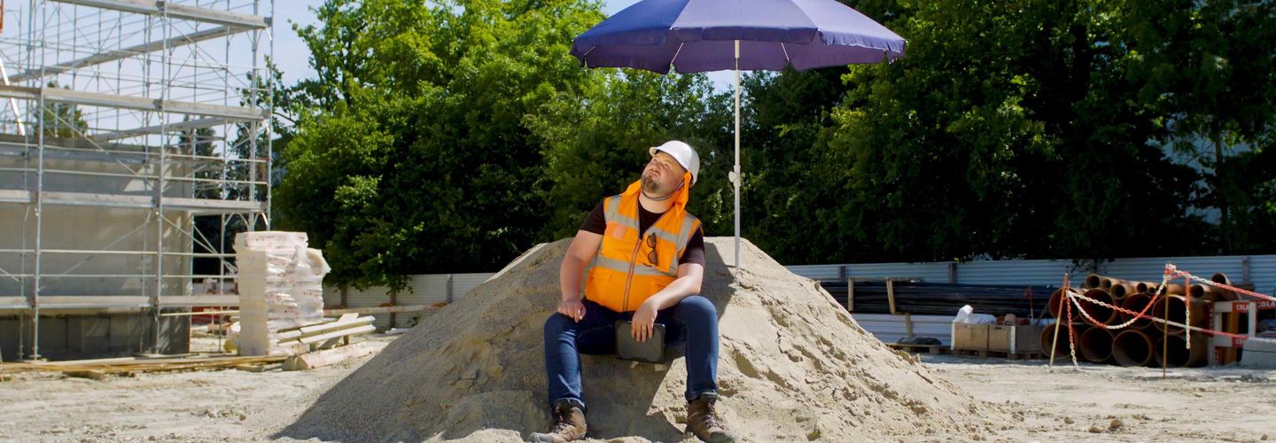 Un homme en gilet orange qui porte un casque de sécurité blanc est assis sur un tas de sable. Il souffre de la chaleur estivale et se met à l’ombre d’un parasol pour fuire les rayons UV. 