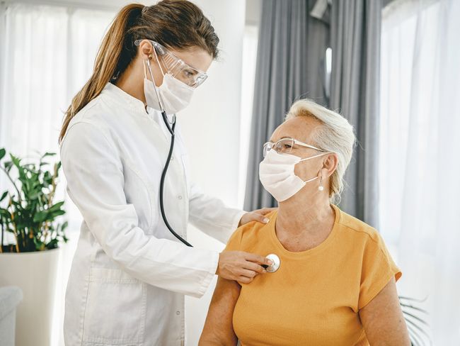 Un médecin ausculte la poitrine d’une patiente. Toutes deux portent un masque de protection respiratoire.