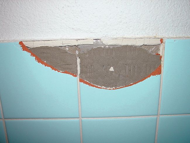 Inquadratura di una parete di un bagno rivestita con piastrelle. È stata staccata una parte di ciascuna delle due piastrelle. Dietro è visibile la colla per piastrelle contenente amianto.