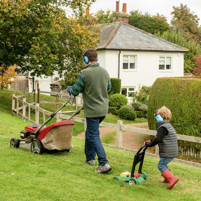 Zuhause, Garten, Rasenmähen, Vater und Sohn, Gehörschutz, Familienzeit, Gartenarbeit, Sicherheit, Outdoor, Wochenendaktivität, Rasenpflege, Natur, Geräuschschutz, Hausarbeit, Eltern-Kind-Moment.