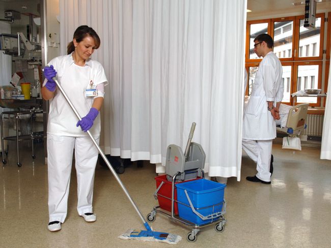 Una donna dai capelli scuri con pantaloni bianchi, casacca bianca e guanti protettivi viola pulisce un pavimento di linoleum. Una tenda bianca la separa da un'area di trattamento medico.