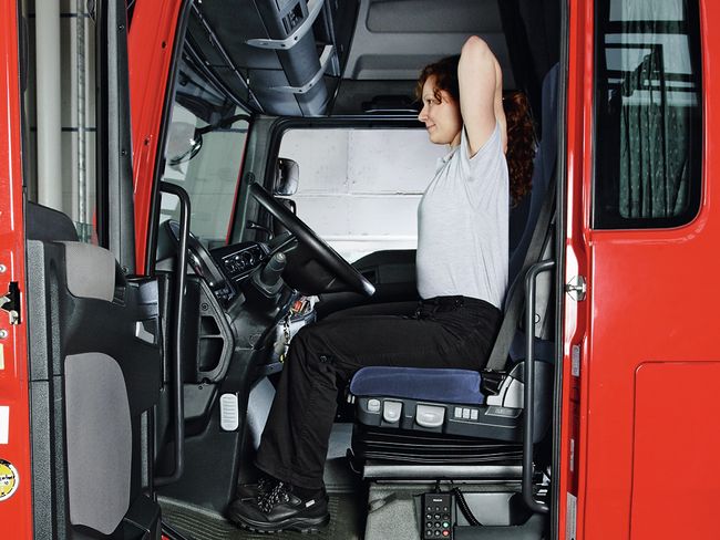 Una donna siede al volante di un camion. Siede con la schiena dritta, appoggia entrambe le mani sulle spalle e porta in alto i gomiti.