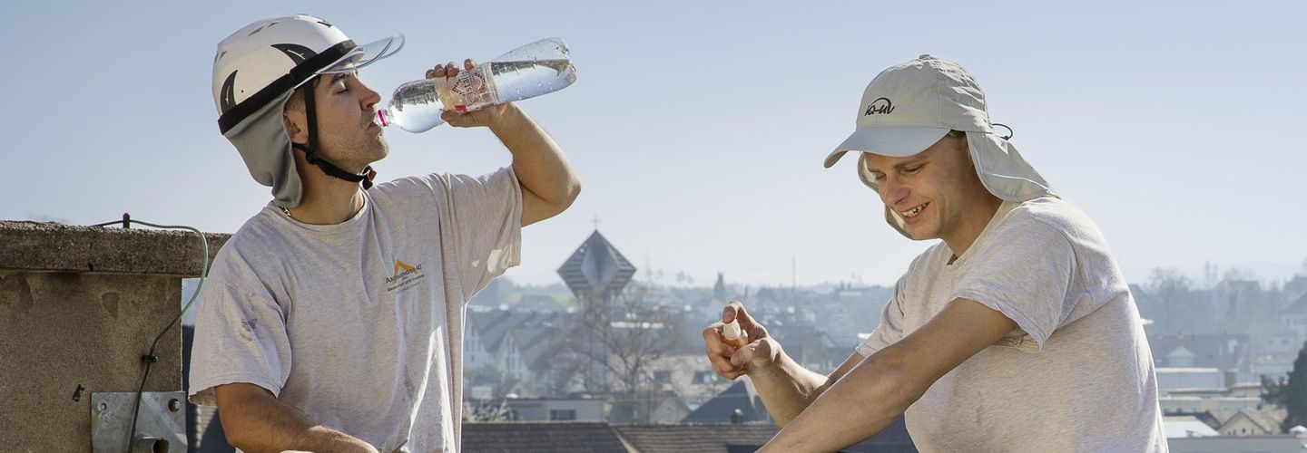 Due carpentieri che indossano pantaloni lunghi scuri, t-shirt bianca e copricapo bianco con visiera e protezione per il collo sono seduti sull’impalcatura di un tetto. La persona a sinistra beve acqua da una bottiglia.
