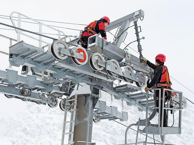 Zwei Personen mit einer persönlichen Schutzausrüstung gegen Absturz reparieren auf einer Seilbahnstütze die Rollenbatterie.