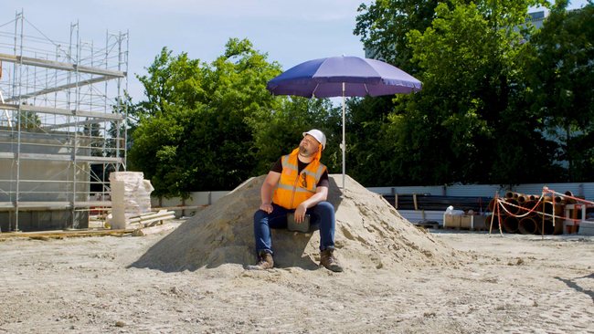 Un homme en gilet orange qui porte un casque de sécurité blanc est assis sur un tas de sable. Il souffre de la chaleur estivale et se met à l’ombre d’un parasol pour fuire les rayons UV. 