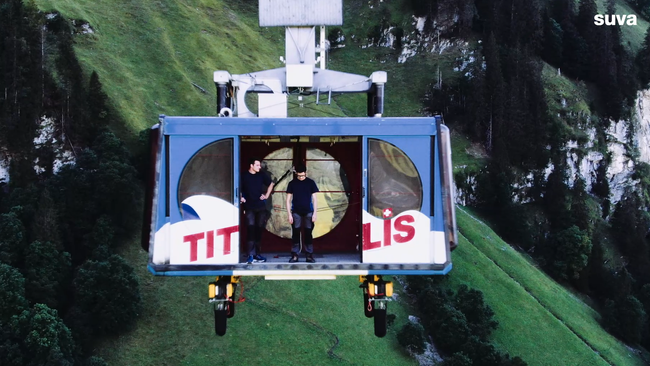 Still aus dem Video. Eine Luftseilbahn schwebt über einem Abgrund. Die Tür steht weit offen. Man sieht zwei junge Männer. Der eine schaut in die Tiefe.