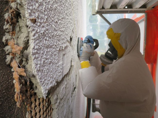Ein Asbestsanierer in einem Schutzanzug und mit einem Atemschutzgerät schleift asbesthaltigen Putz ab.