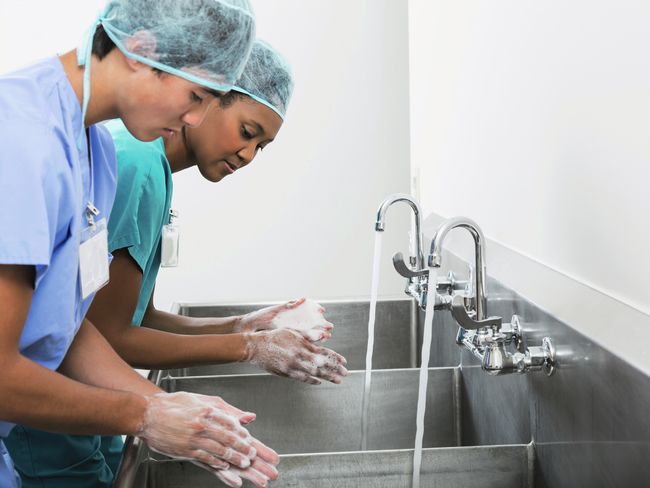 Zwei Pflegekräfte in grüner und blauer Schutzkleidung und grünem Haarschutz schäumen sich die Hände an zwei Aluminiumbecken unter laufendem Wasser ein.