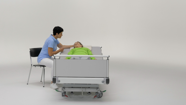 On voit une femme couchée sur le dos sur un lit médicalisé. Une personne aidante est assise sur une chaise à côté d’elle. Le lit est réglé pour supporter son propre poids lors des soins, en s’appuyant sur un bras.