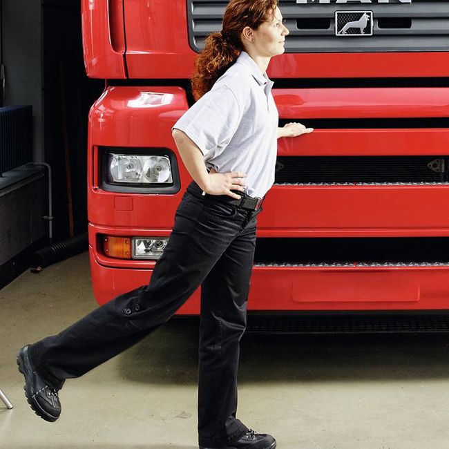 Une femme se tient de profil devant un camion. Avec la main gauche, elle s’appuie légèrement sur le véhicule. Sa main droite se trouve au niveau de la taille et sa jambe droite se balance vers l’arrière.