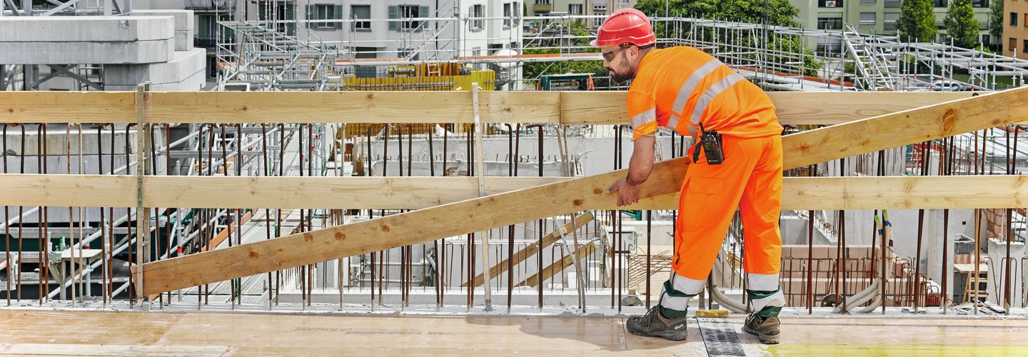 Ein Mann auf einer Baustelle mit Sicherheitsausrüstung hält ein langes Brett vor einem Geländer und steht leicht gebückt.