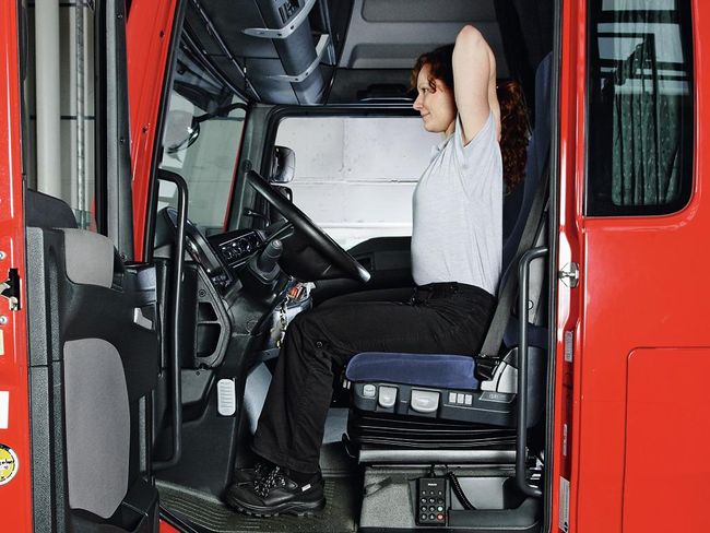 Una donna siede al volante di un camion. Siede con la schiena dritta, appoggia entrambe le mani sulle spalle e porta in alto i gomiti.