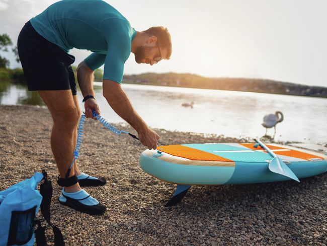 Ein junger Mann pumpt mit einer Handluftpumpe ein Standup-Paddleboard am Ufer eines Sees auf.