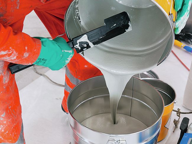 Un operaio che indossa una tuta protettiva arancione e guanti verdi versa un liquido chiaro e viscoso da un secchio in un secchio posto più in basso contenente una massa di colore analogo.