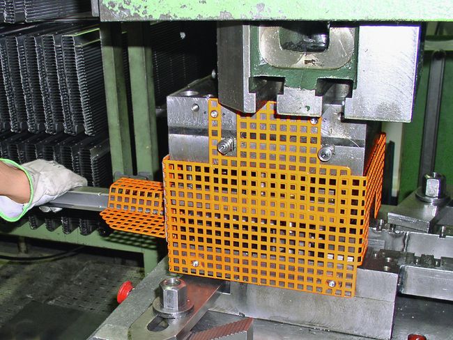 Une grille métallique orange entoure une presse. Un travailleur introduit un profilé métallique dans la presse par la gauche.