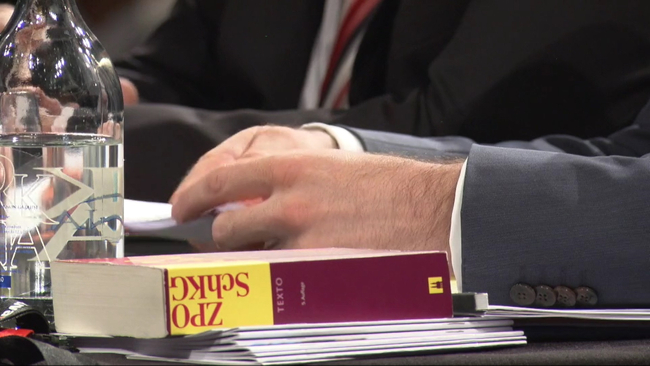 Ein Buch über die Zivilprozessordnung (ZPO) liegt auf einem Tisch. Daneben ist die Hand eines Richters und ein Glas Wasser zu sehen. 