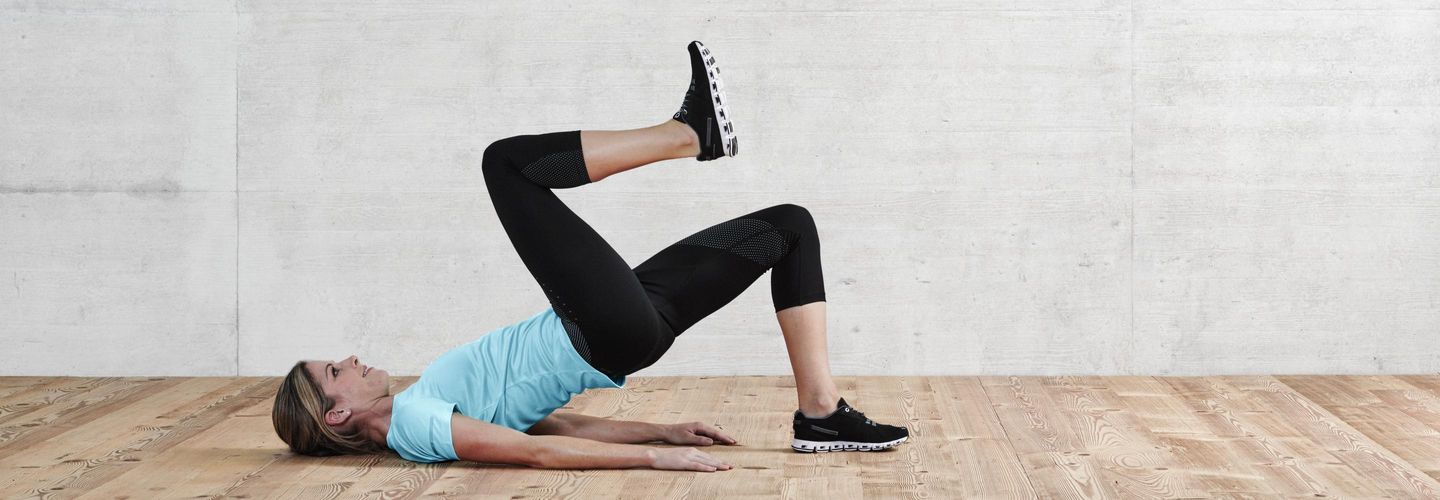 Eine Frau in Sportkleidung ist in der Schulterbrücke. Sie hebt ihr rechtes Knie in Richtung Brust.