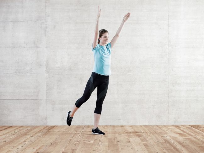 Eine Frau in Sportkleidung macht eine Fitnessübung. Sie steht auf dem linken Bein, streckt das rechte Bein gestreckt nach hinten und reckt die Arme in die Höhe.
