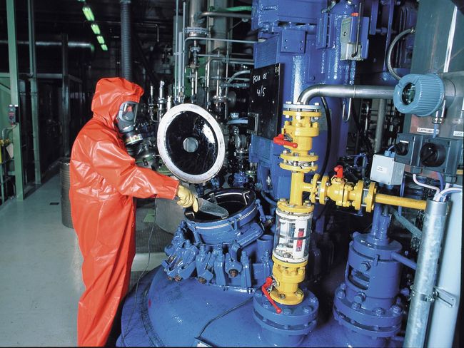 Fachkraft in Chemikalienschutzkleidung mit Atemschutz-Vollmaske entnimmt Probe aus Tank