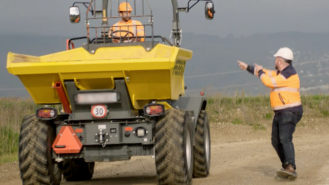 Un homme en pull orange montre des doigts une machine de chantier qui passe à côté de lui. 