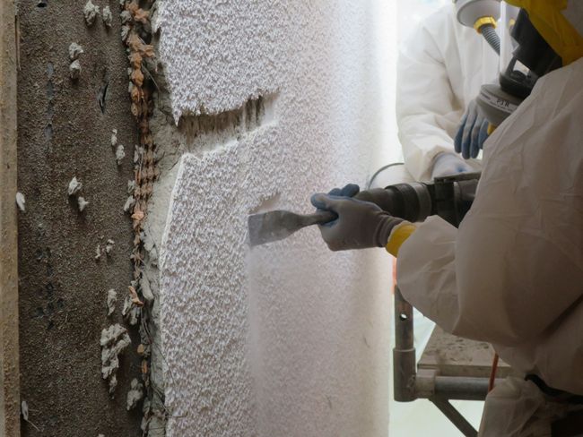 Ein Asbestsanierer in einem Ganzkörperschutzanzug und mit einem Atemschutzgerät spitzt den asbesthaltigen Putz mithilfe eines Elektroschabers vom Untergrund.
