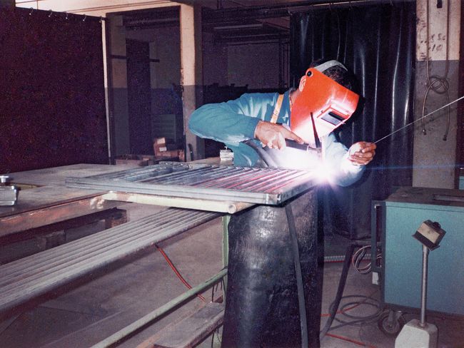 Ein Handwerker mit einer Schweissmaske arbeitet an einem Werkstück. Der Gesichtsschutz bedeckt auch den Halsbereich, das dunkle Sichtfenster schützt die Augen vor Partikeln und dem grellen Licht.