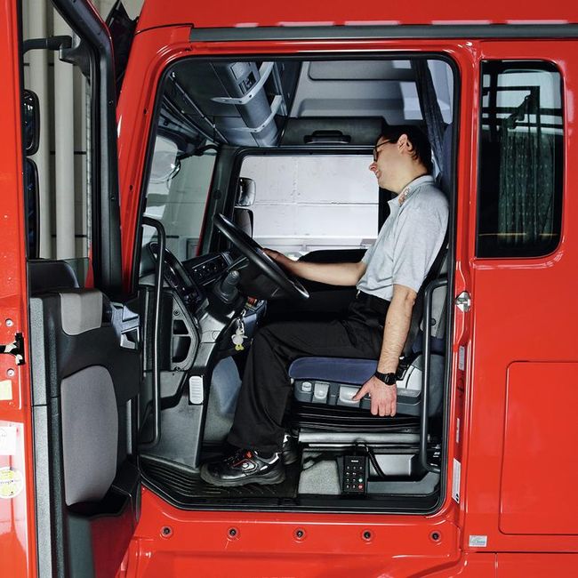 Un uomo siede al volante di un camion. Con la mano destra si tiene rilassato al volante, con la mano sinistra al sedile. Guarda in avanti e piega la testa verso destra.