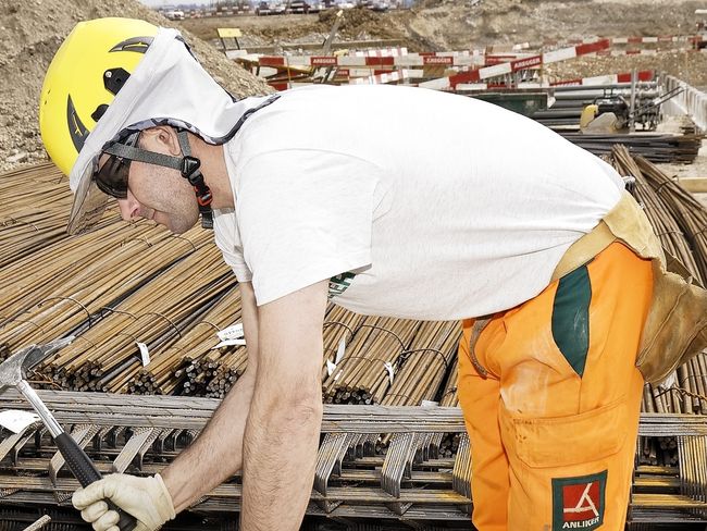 Bauarbeiter beugt sich über seine Arbeit auf einer Baustelle. Er trägt eine orange Hose, ein weisses T-Shirt und einen gelben Helm mit Stirnblende und weissem Nackenschutz.