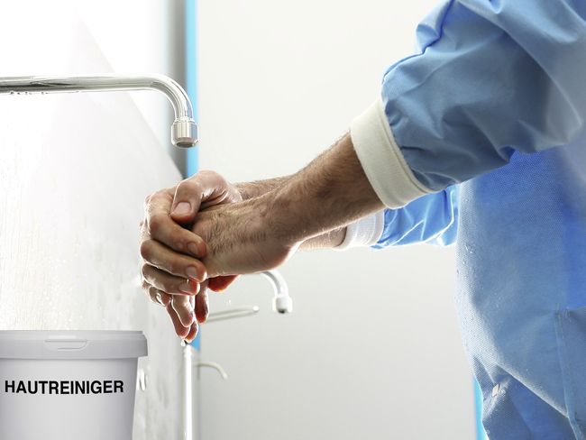 Männliche Person wäscht die Hände unter einem Wasserhahn. Vor ihm steht ein Behälter mit der Aufschrift «Hautreiniger».