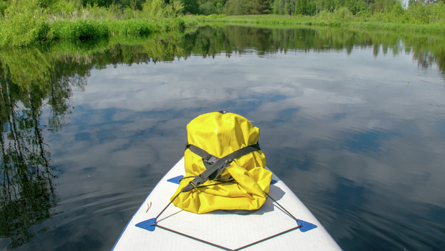 Sommerlandschaft am Fluss, im Vordergrund ein SUP-Board und ein gelber Gummibeutel, mit Wolken, die sich im Wasser spiegeln, sowie grünen Bäumen und Gras am Ufer.